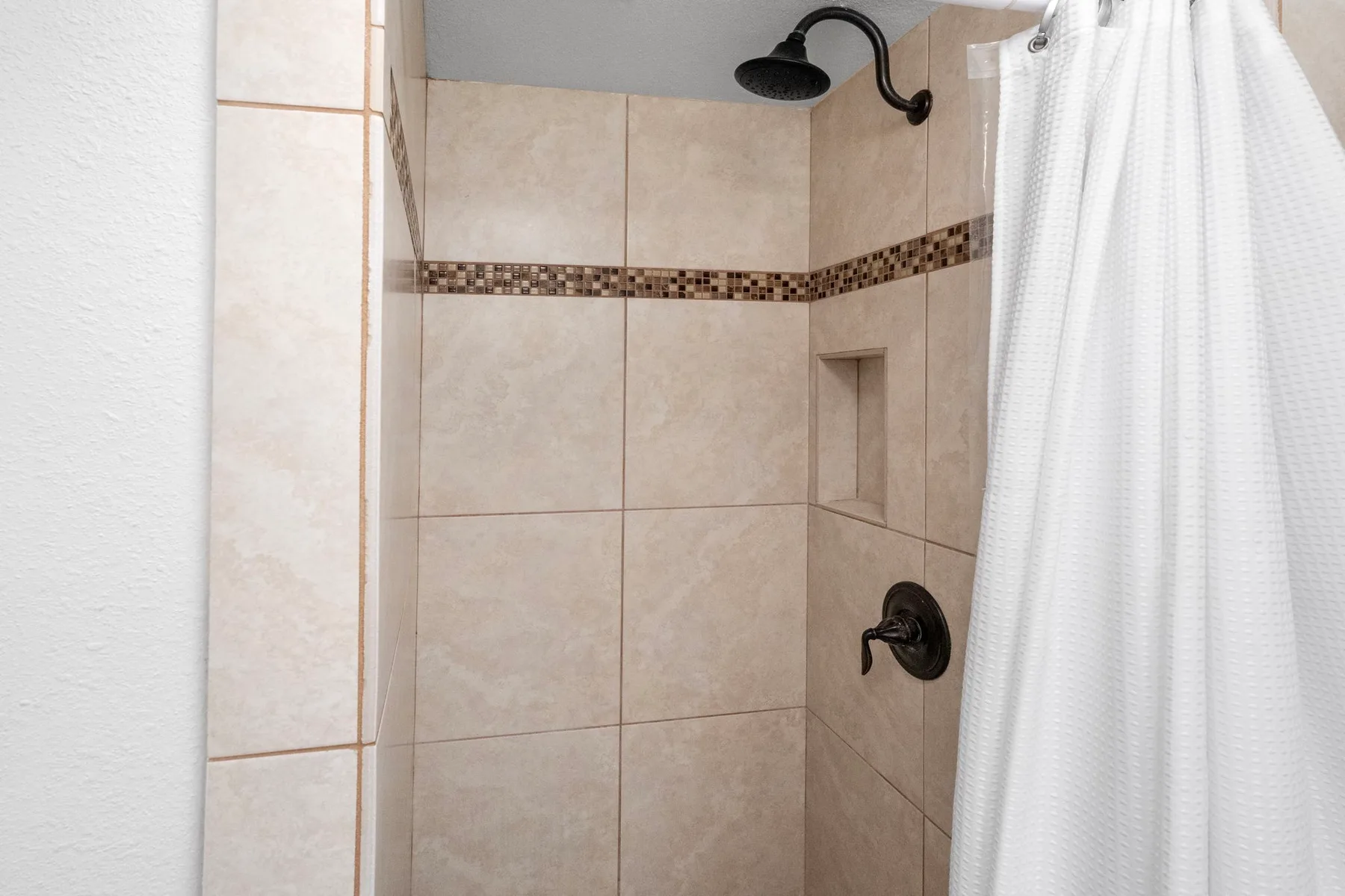Una ducha con azulejos, un cabezal oscuro y una cortina blanca.
