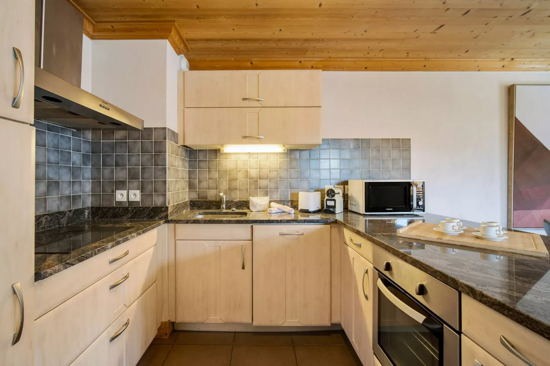 Una cocina compacta con gabinetes de madera clara, encimeras oscuras y electrodomésticos de acero inoxidable.