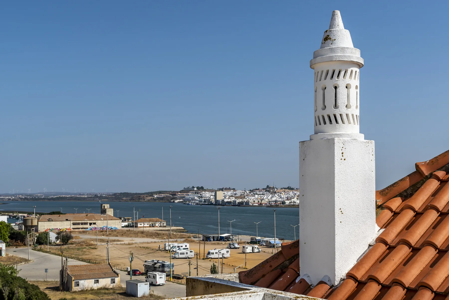 Una chimenea blanca sobresale sobre tejas de terracota, con un pueblo costero al fondo.