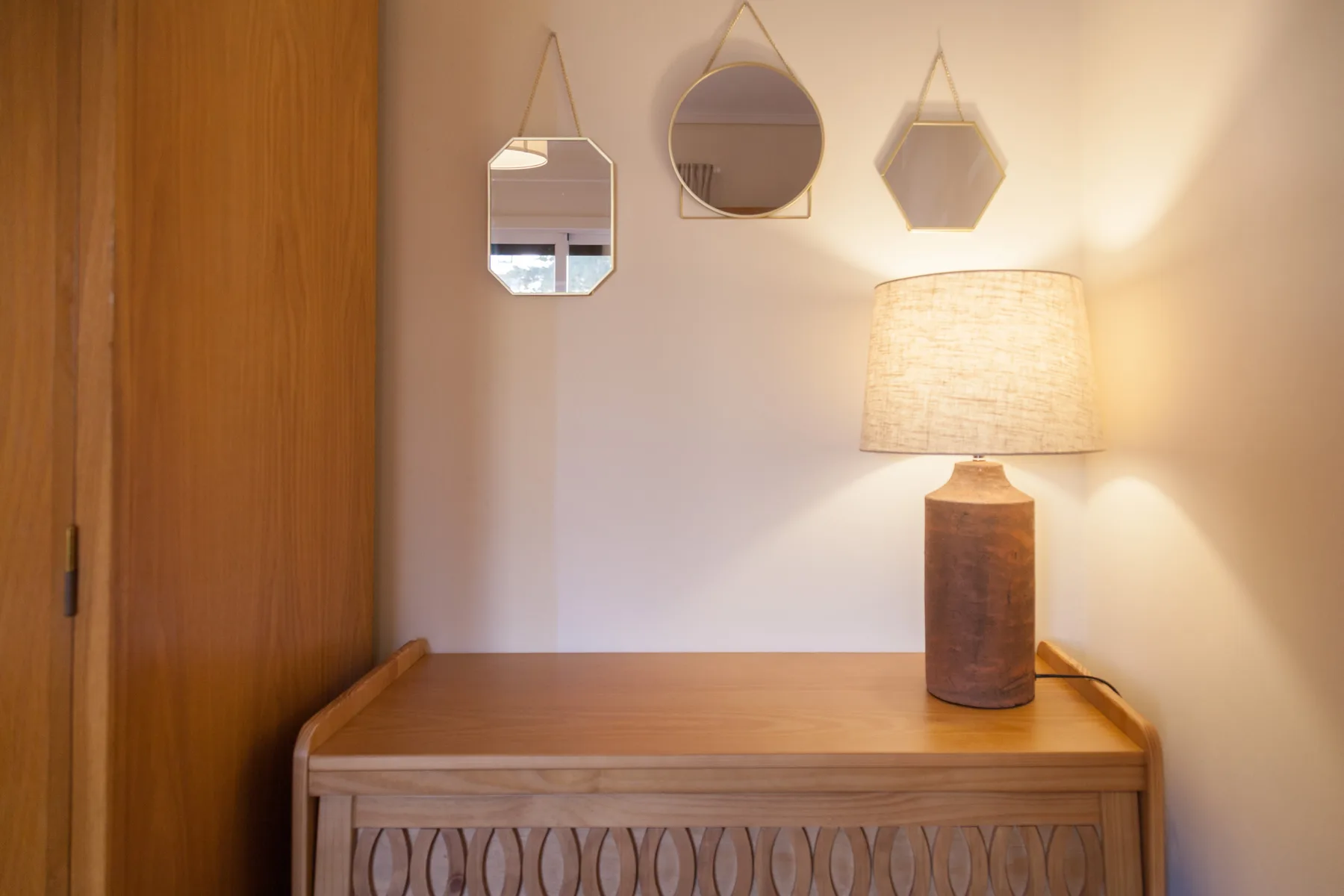 Una consola de madera está bajo tres espejos decorativos, junto a una lámpara encendida.