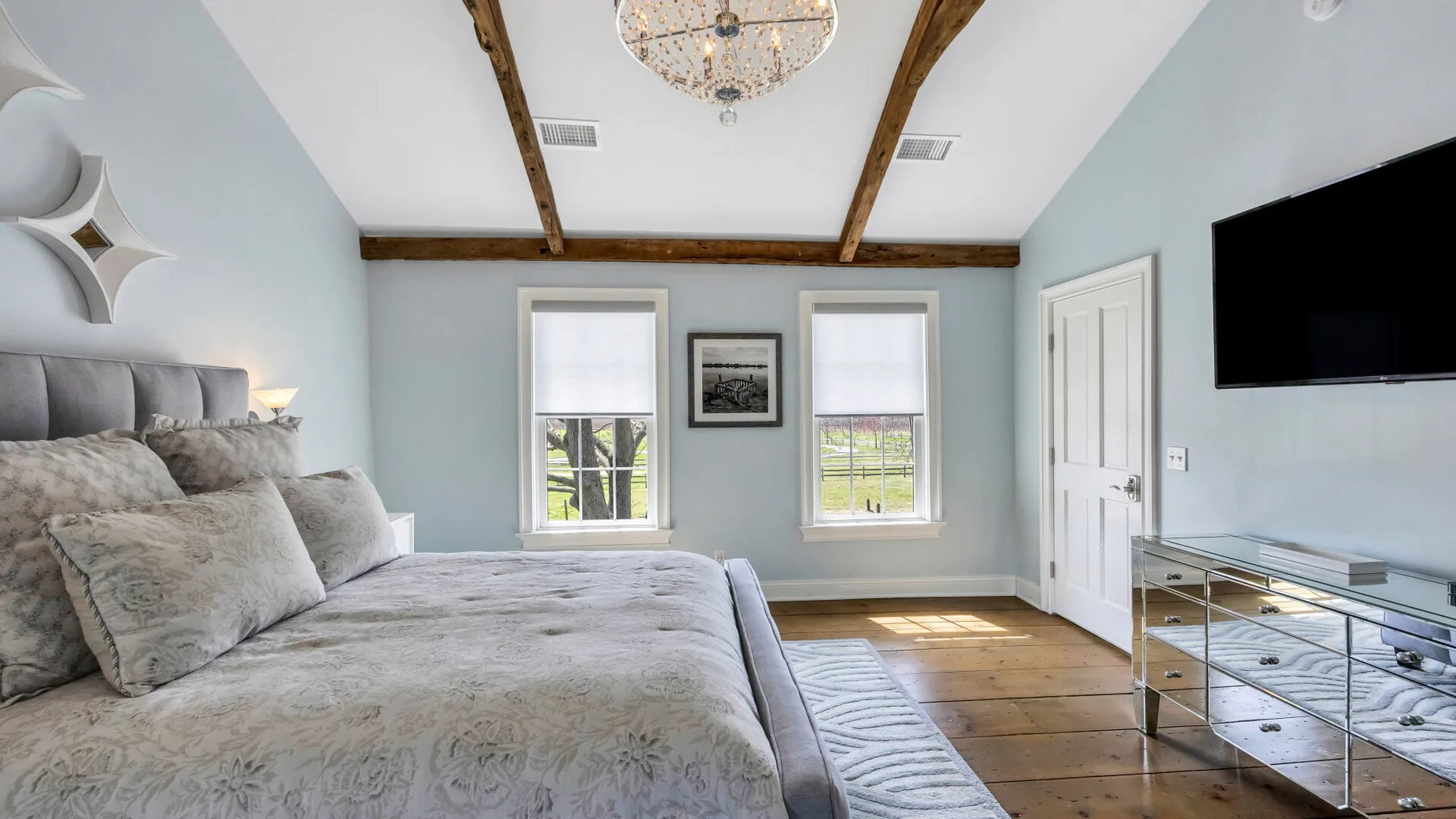 Un dormitorio luminoso con una cama grande, dos ventanas y un televisor en la pared.