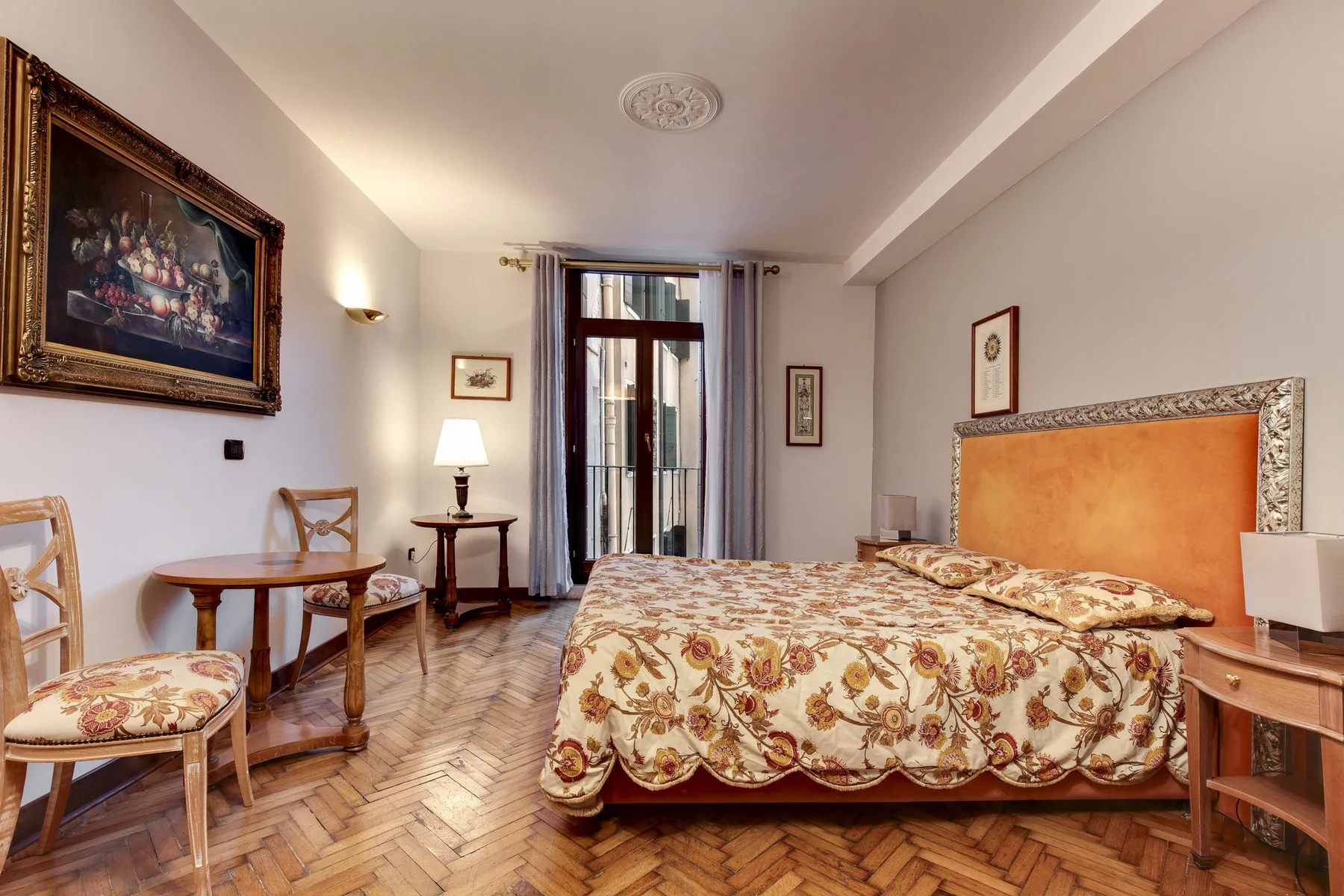 Un dormitorio con una colcha floral, asientos y una ventana alta con cortinas.