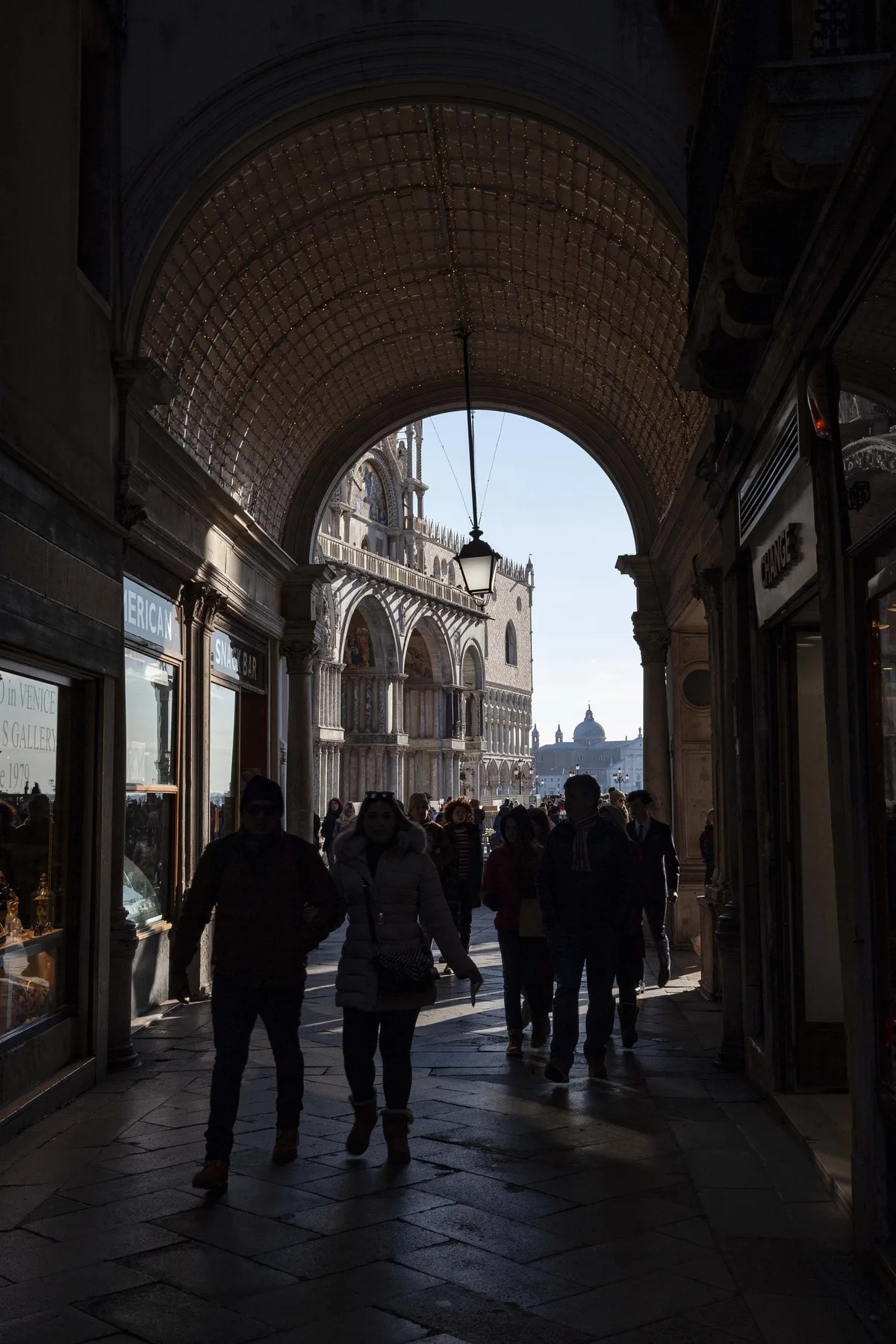 Una galería cubierta da a una plaza histórica, mientras pasan peatones.