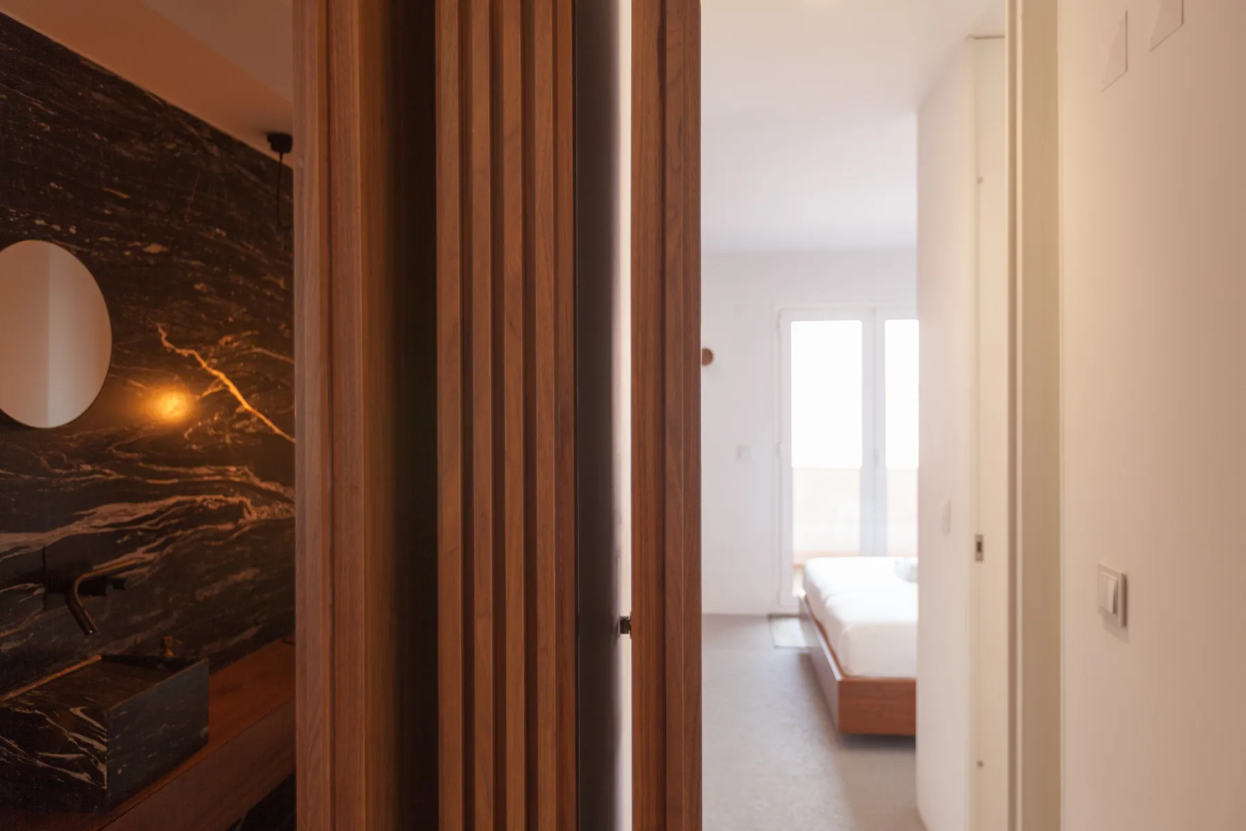 Se ve un dormitorio a través de una puerta del pasillo, con una cama junto a una ventana luminosa.