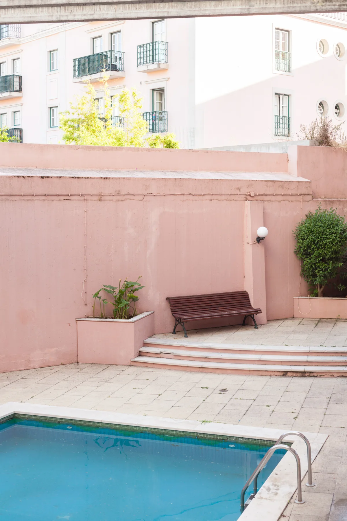 Zona de piscina exterior con suelo de baldosas y un banco junto a una pared rosa.