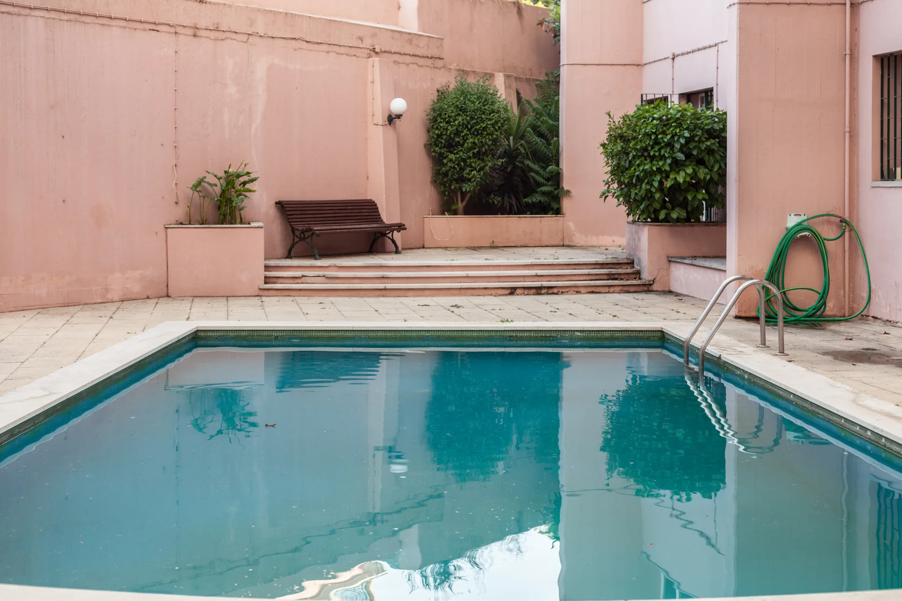 Una piscina al aire libre está en un patio con paredes rosadas y plantas en macetas.