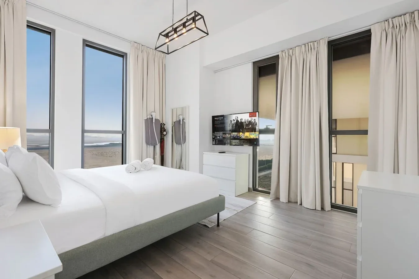 Un dormitorio luminoso con una cama tendida, ventanas altas y un televisor.