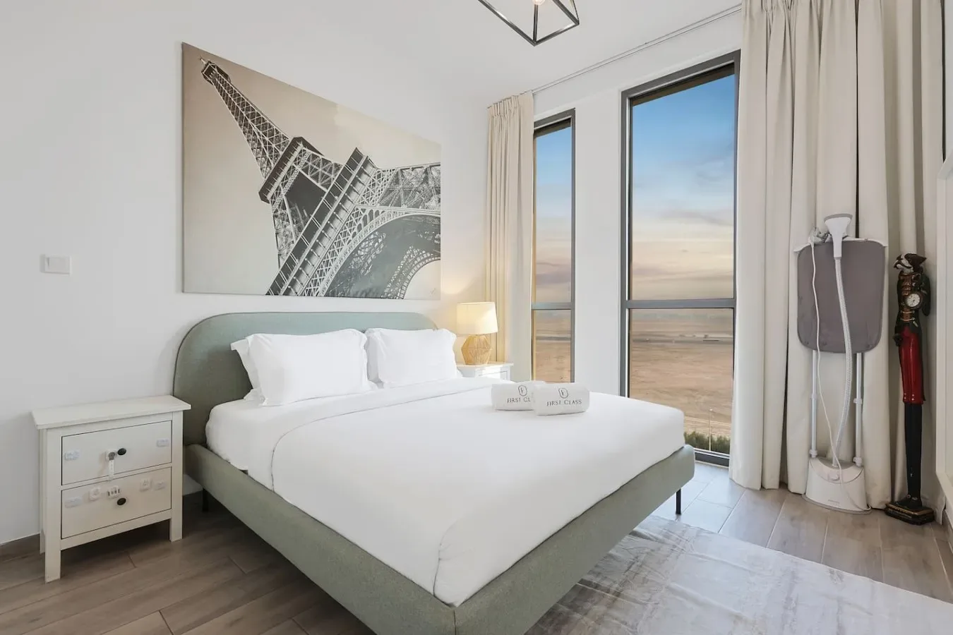 Un dormitorio con una cama bien hecha junto a ventanas altas y cortinas claras.