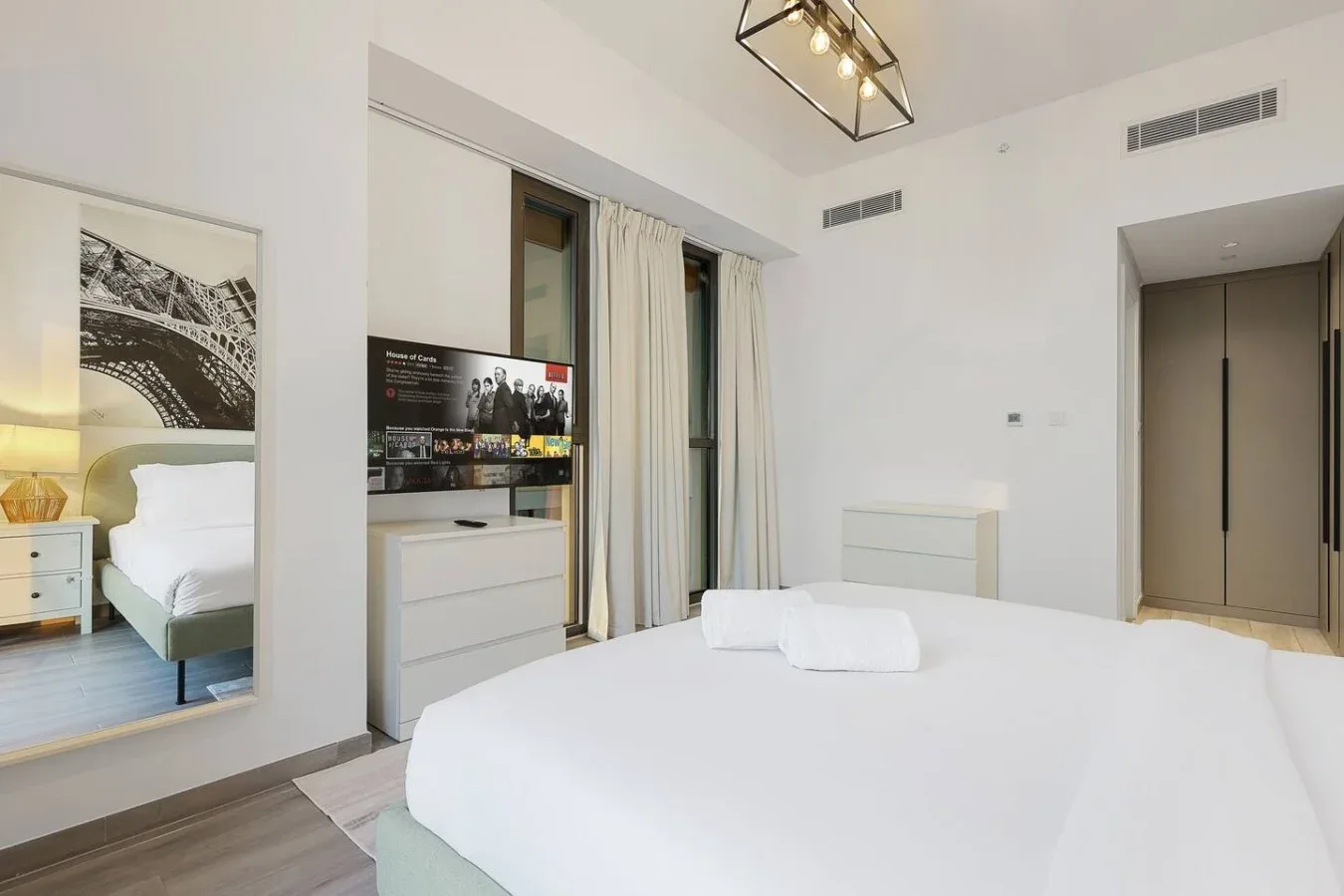 Un dormitorio luminoso con una cama blanca, televisión y cortinas largas.