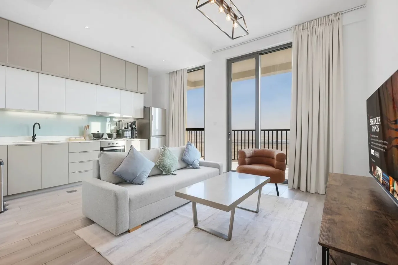 Una luminosa sala de estar de planta abierta tiene una zona de asientos junto a una cocina moderna y ventanas altas.