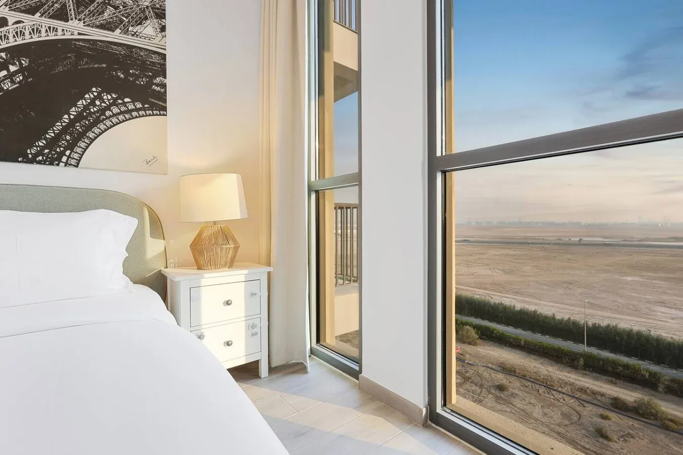 Un dormitorio con una cama blanca junto a ventanas altas con vistas a un paisaje abierto.