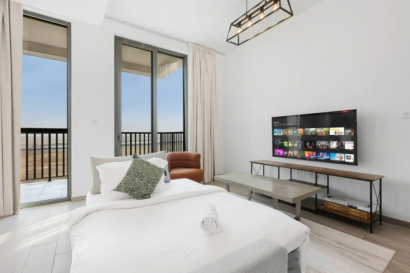 Un dormitorio luminoso con una cama blanca, grandes ventanales y un televisor.