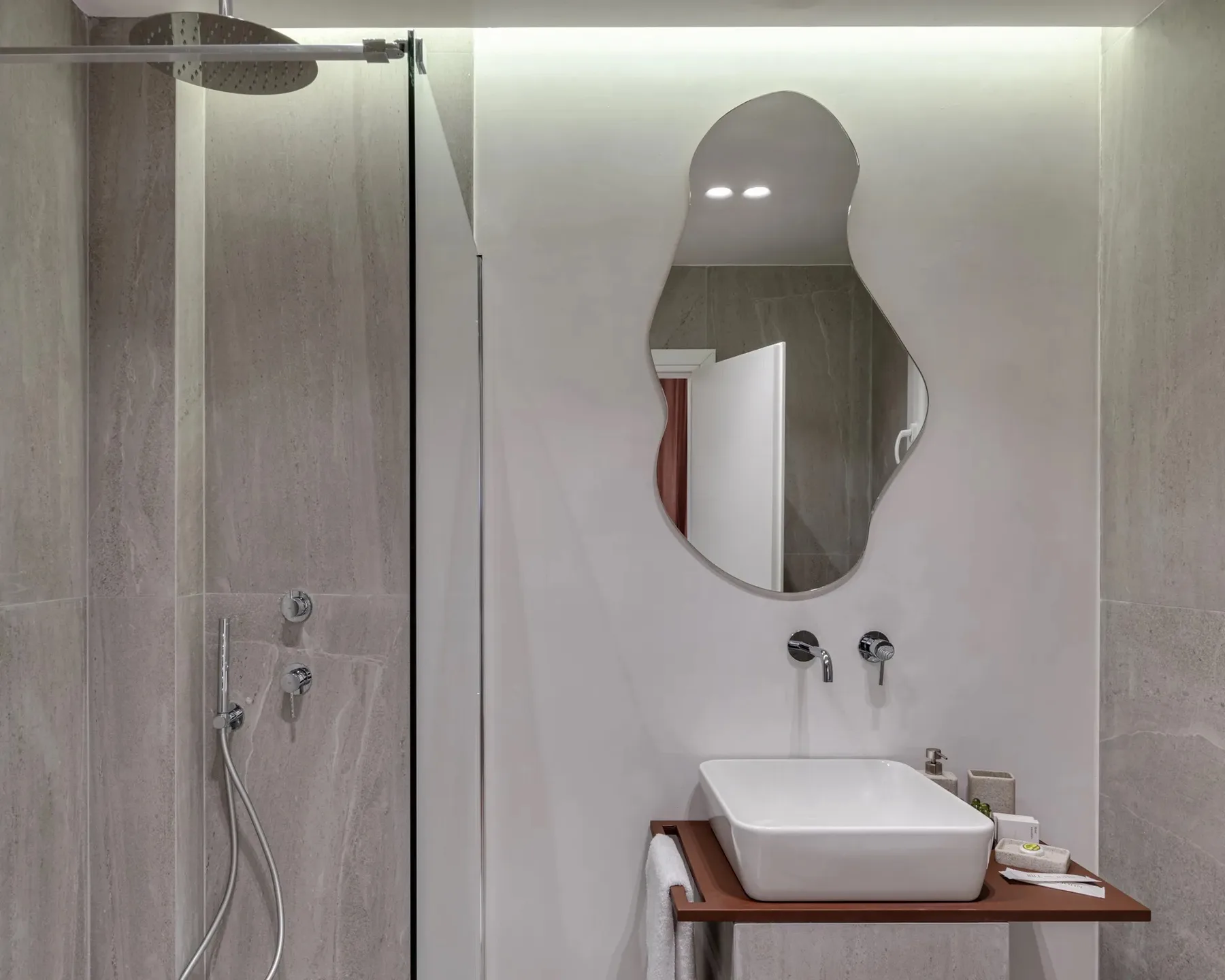 Un baño moderno con ducha y lavabo bajo un espejo de forma irregular.