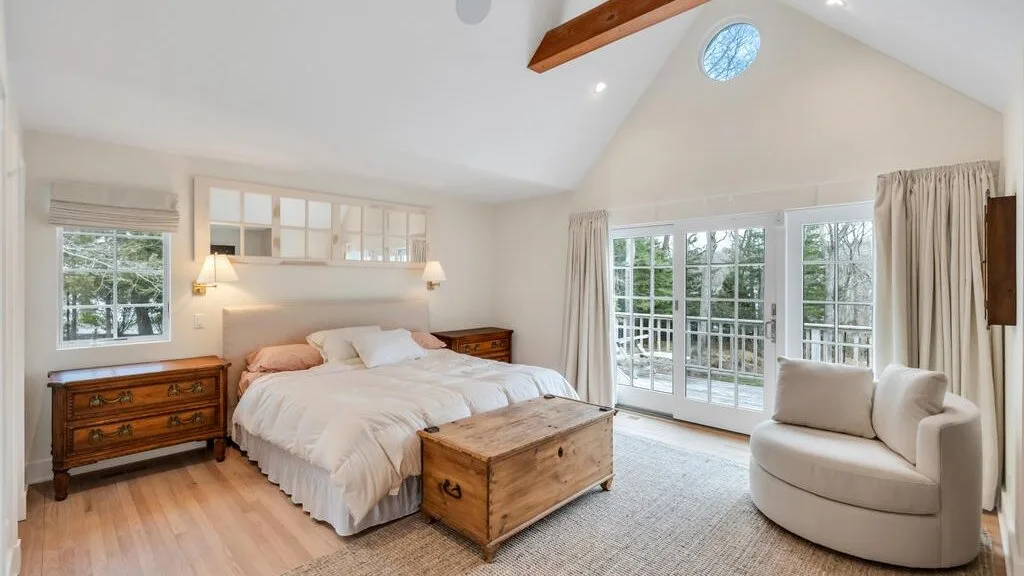 Un dormitorio luminoso con una cama, un arcón de madera y ventanales.