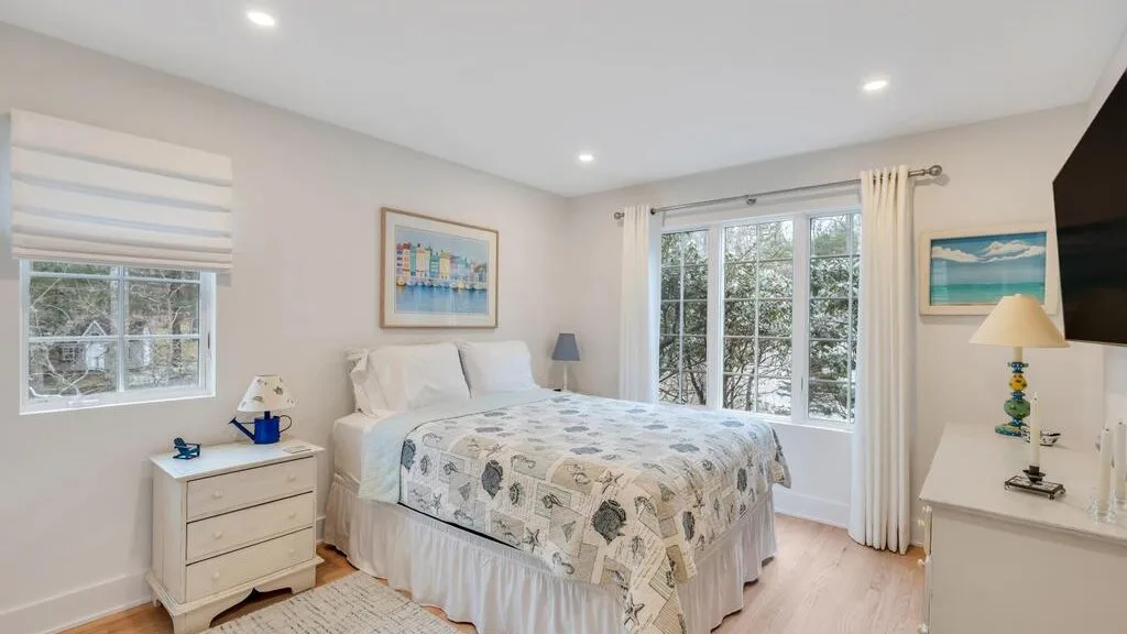 Un dormitorio luminoso con una cama junto a grandes ventanas con vistas a los árboles.