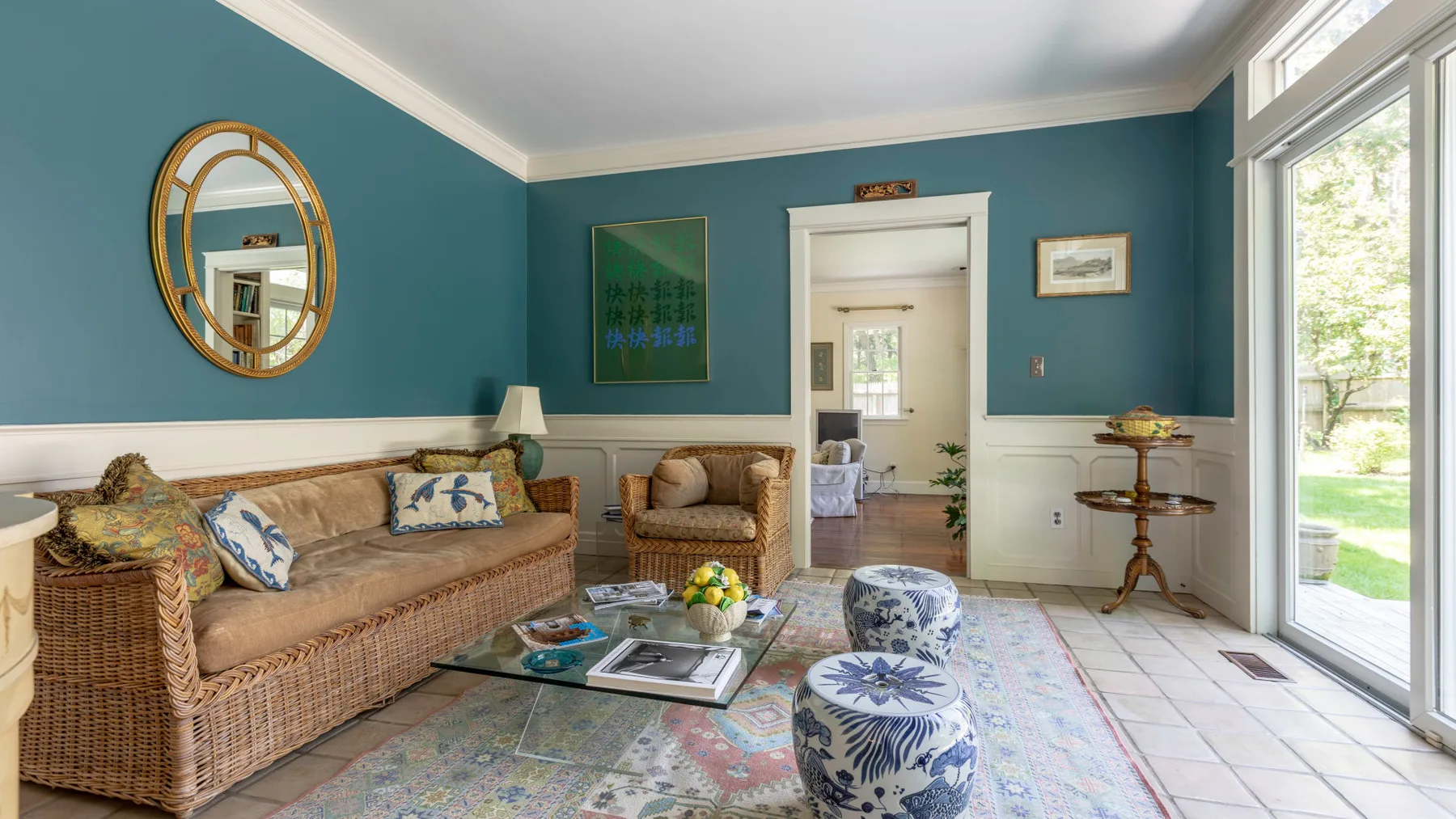 Una sala de estar color verde azulado con asientos de mimbre, una alfombra estampada y grandes ventanales.