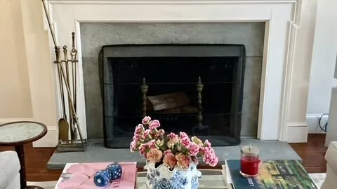 Una chimenea en una sala de estar con un jarrón de flores rosadas en el hogar.