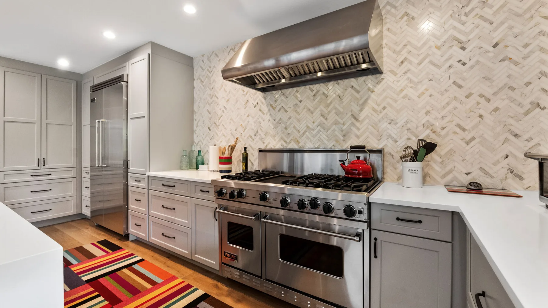 A bright kitchen with gray cabinetry, stainless steel appliances, and a colorful striped rug.