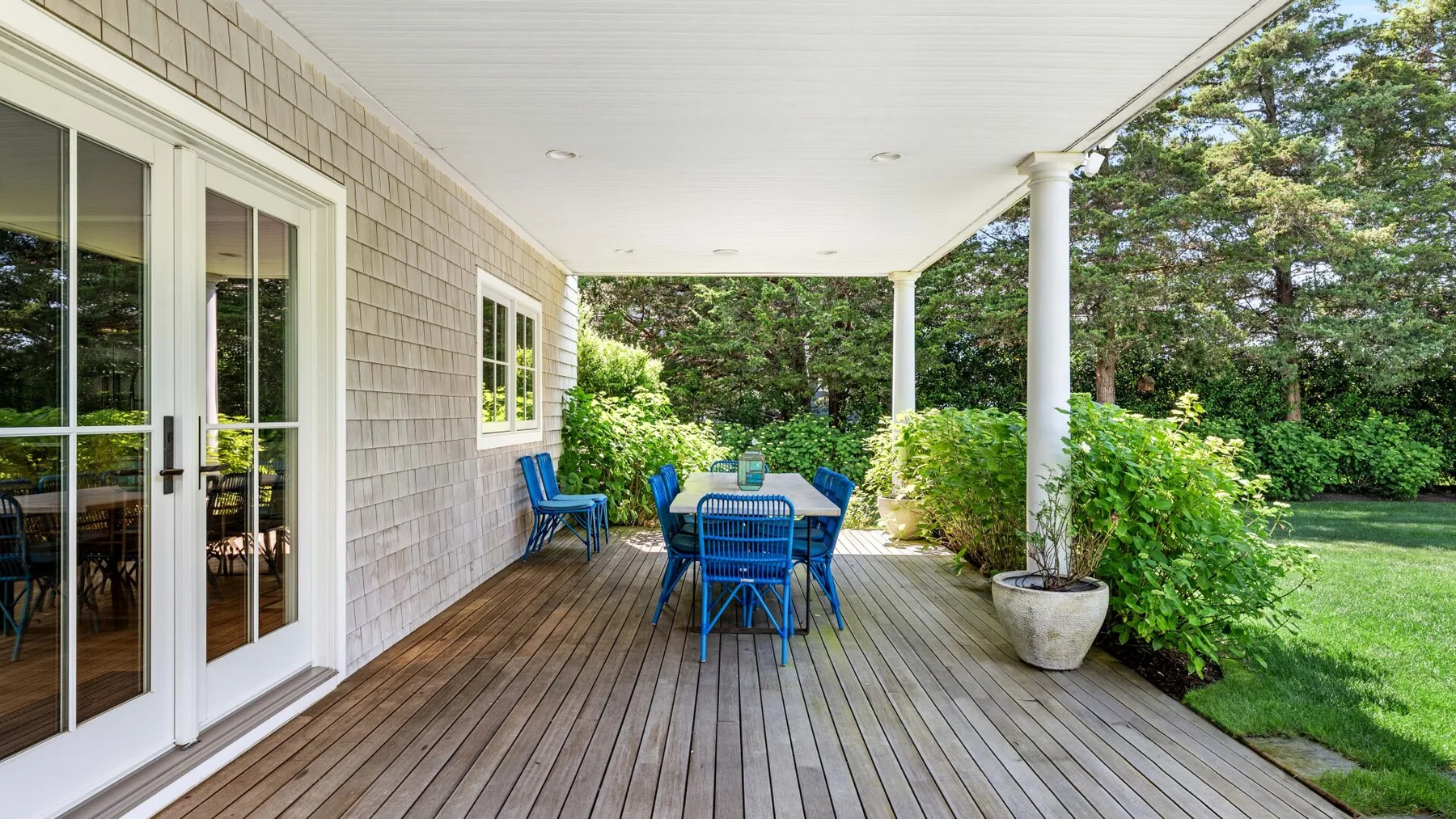 A covered outdoor deck with blue seating and a table overlooks a green yard.