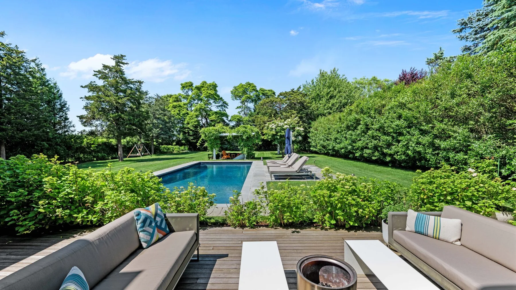 Outdoor seating overlooks a swimming pool surrounded by green hedges and trees.