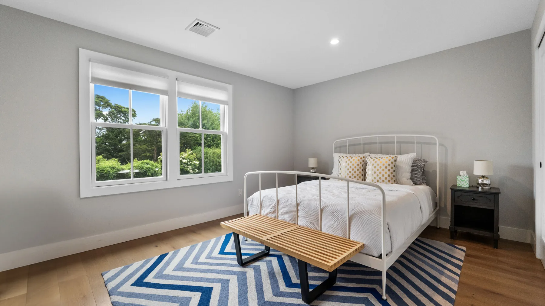 A bedroom with a bed, a striped rug, and windows overlooking greenery.