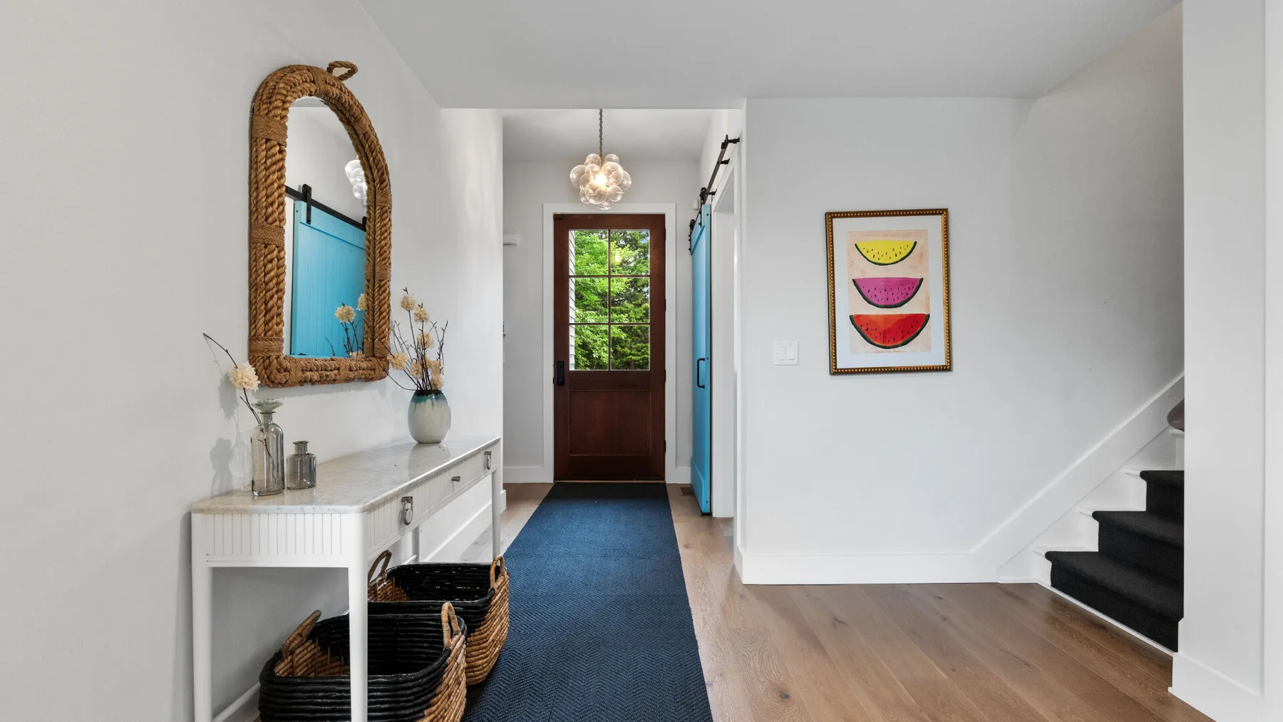 A bright entry hallway with a console table, woven mirror, and dark staircase.