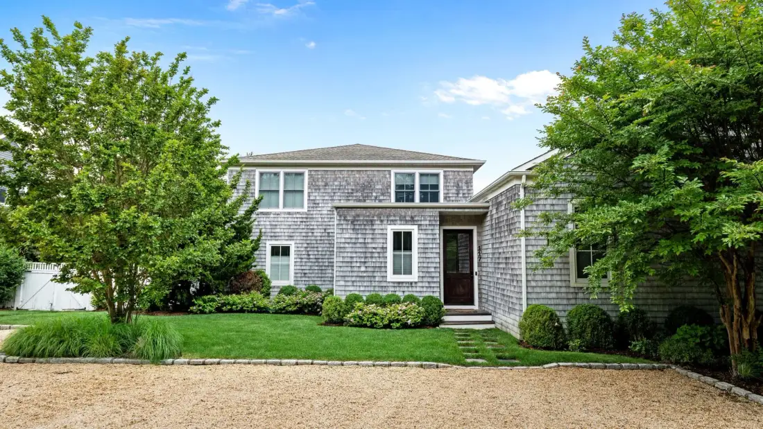 A shingle-sided house stands behind a lawn and gravel driveway.