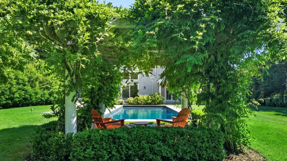 A garden patio with a swimming pool and outdoor seating beneath leafy trees.