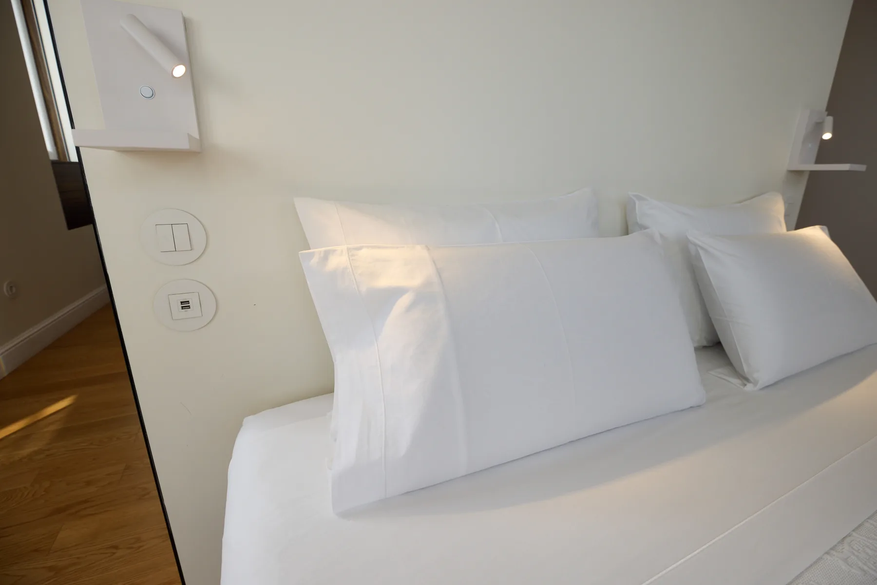 A white bed with layered pillows sits beside a light-colored wall.