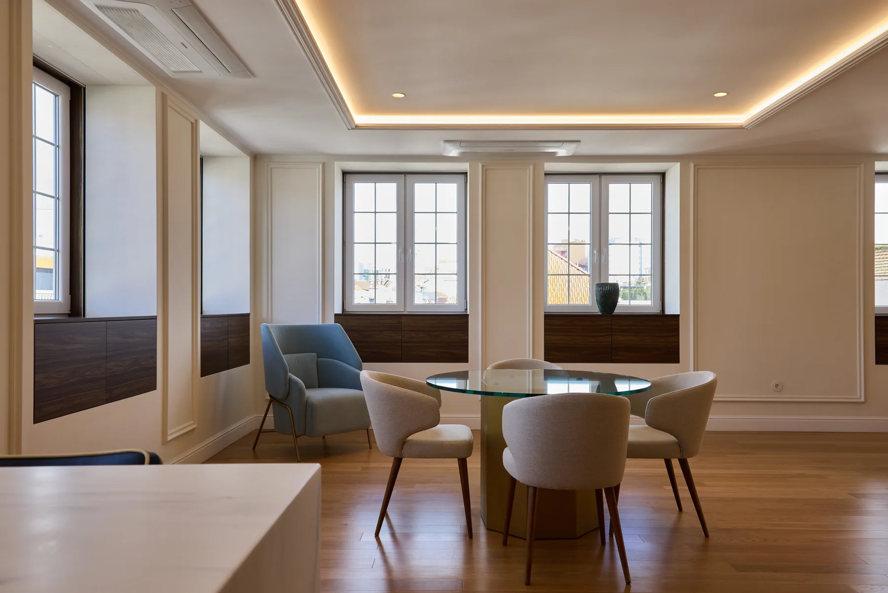 A dining area with a round table and upholstered seating sits beside large windows.