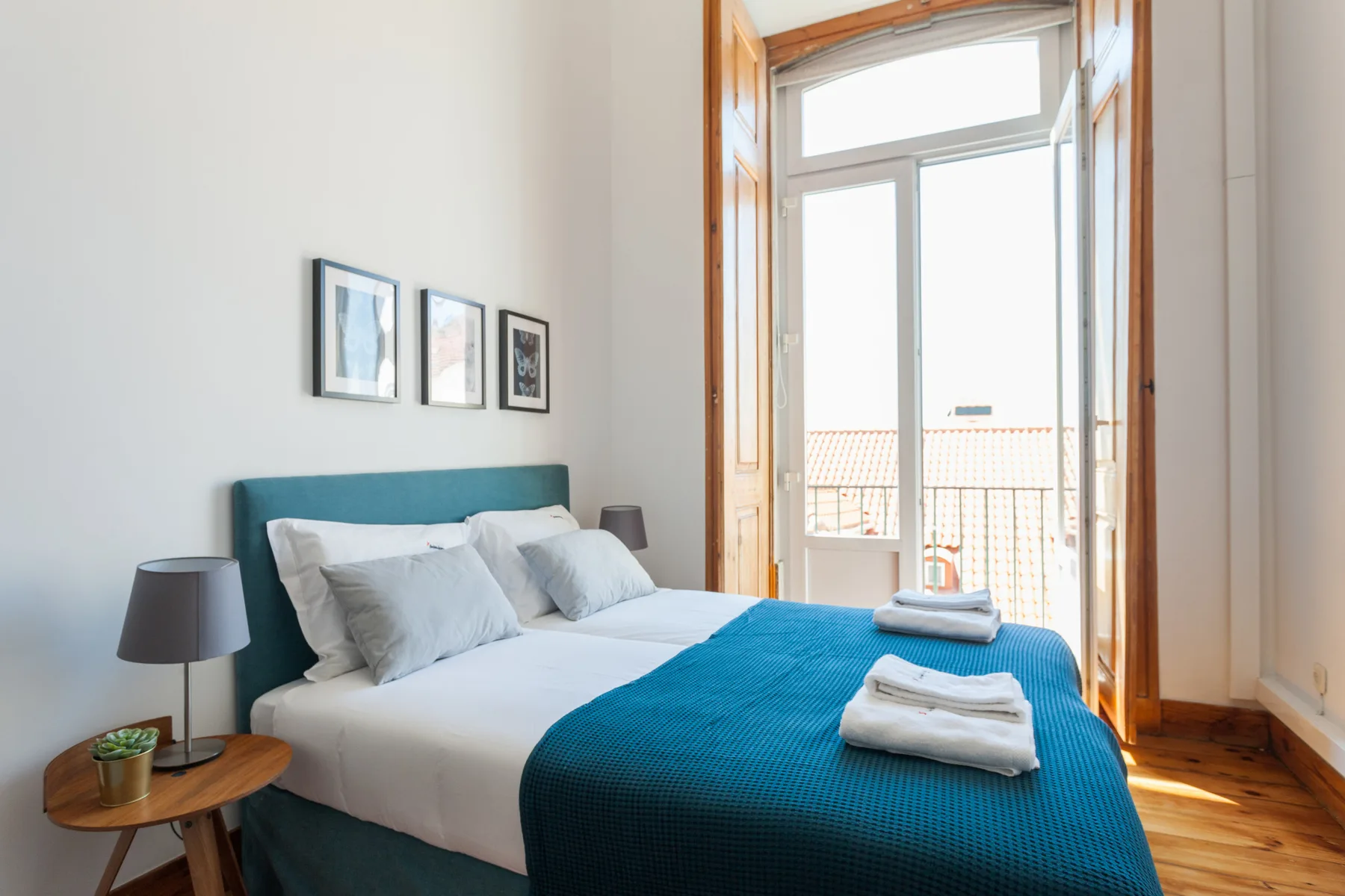 A bright bedroom with a bed, framed pictures, and an open balcony door.