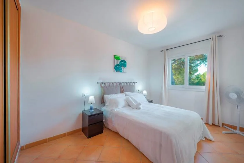 A bright bedroom with a neatly made bed and a window overlooking greenery.