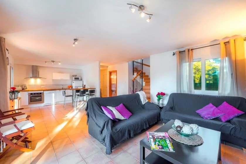 A bright open-plan living room with dark seating beside a kitchen and dining counter.