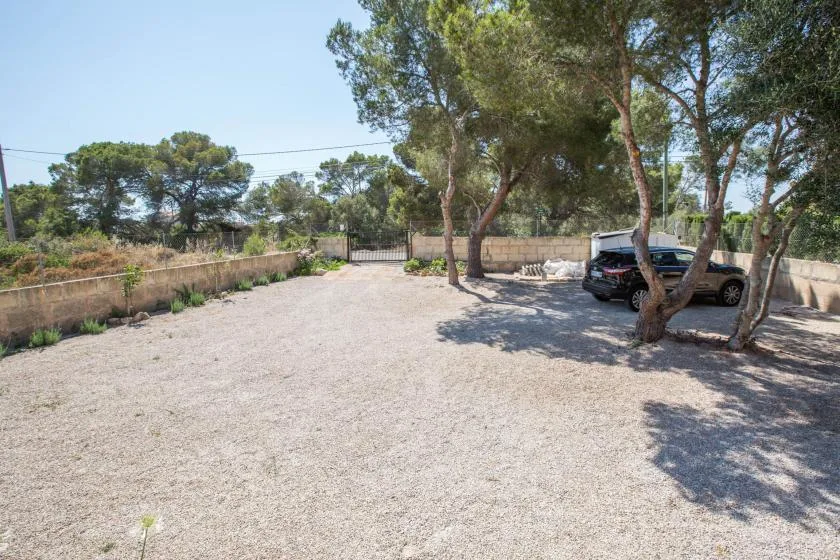 A gravel parking area bordered by trees, low walls, and a parked car.