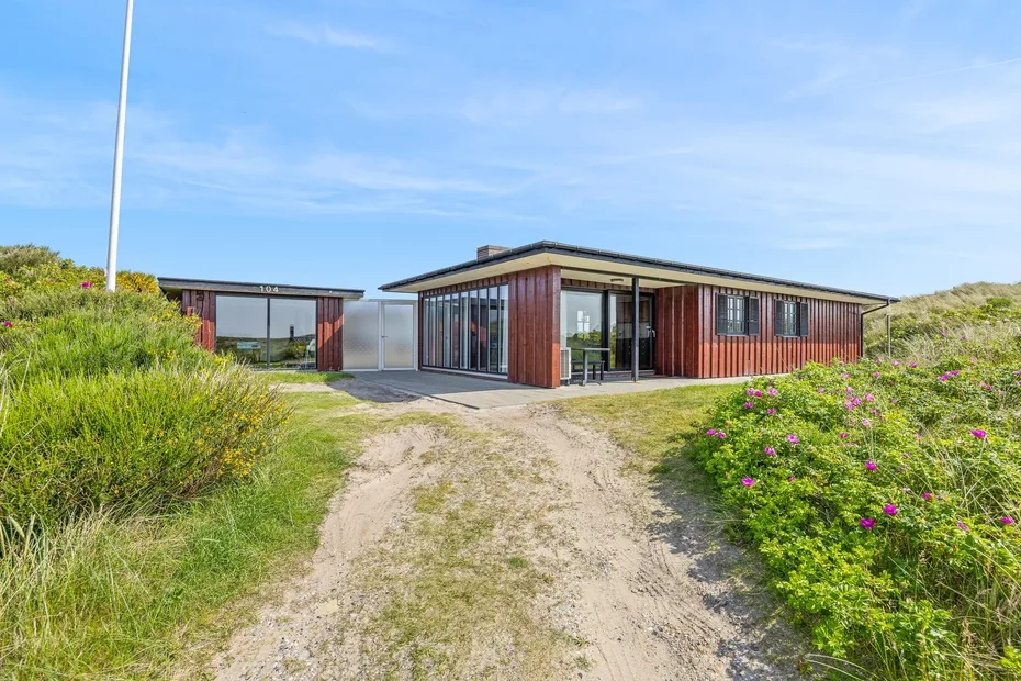 A modern building with broad windows stands beside a sandy path and flowering greenery.