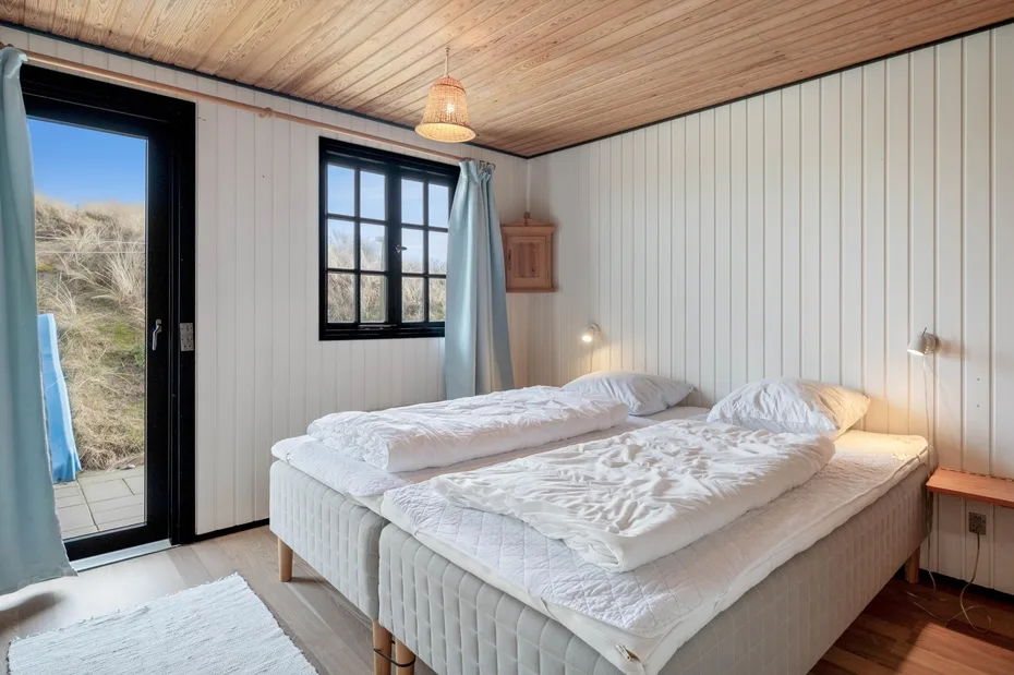 A bedroom with a large bed, white bedding, and windows overlooking grassy dunes.