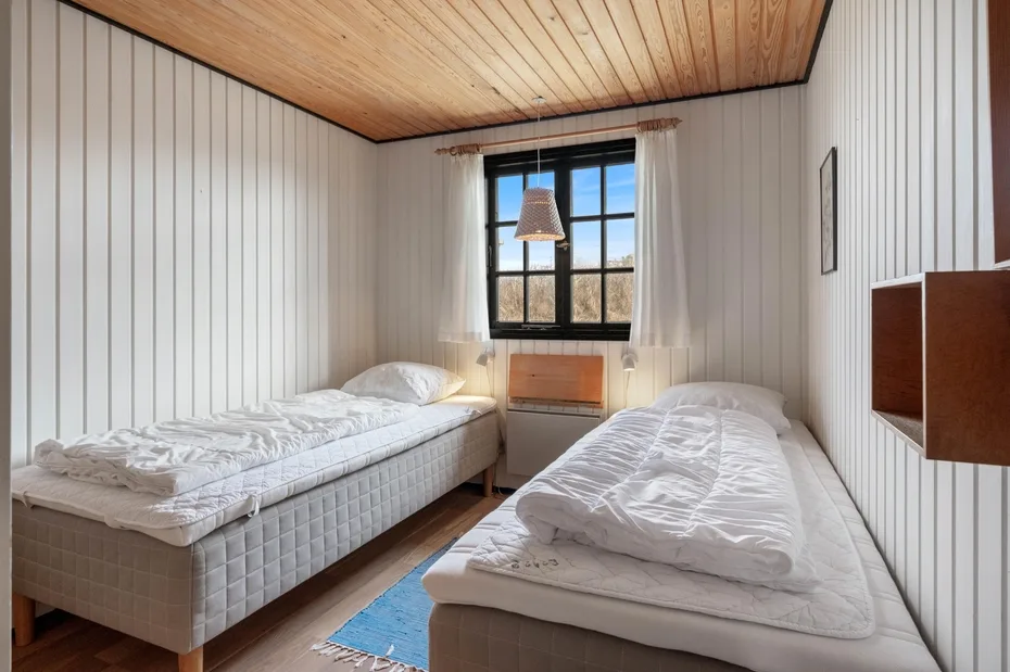 A bedroom with two beds, a window, and white paneled walls.