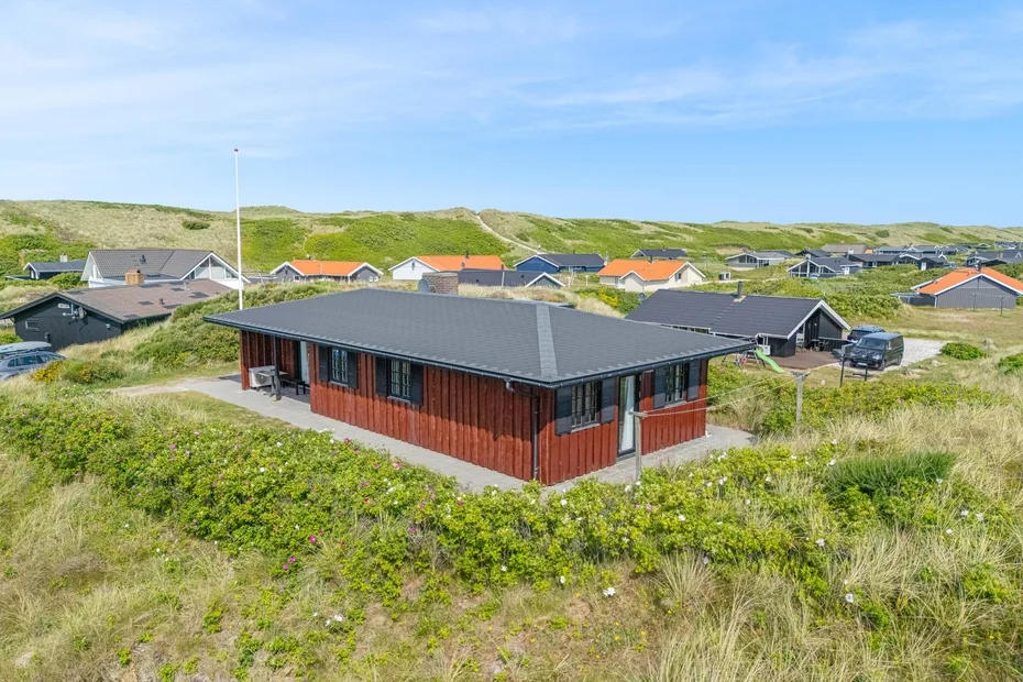 A red-sided house sits among grassy dunes and other homes.