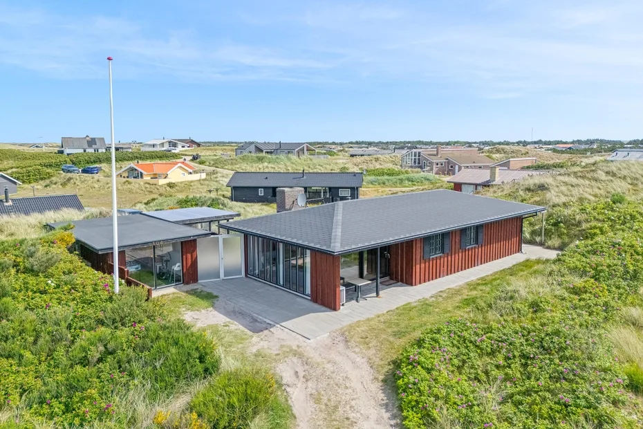A modern house sits among grassy dunes, with neighboring homes visible in the distance.