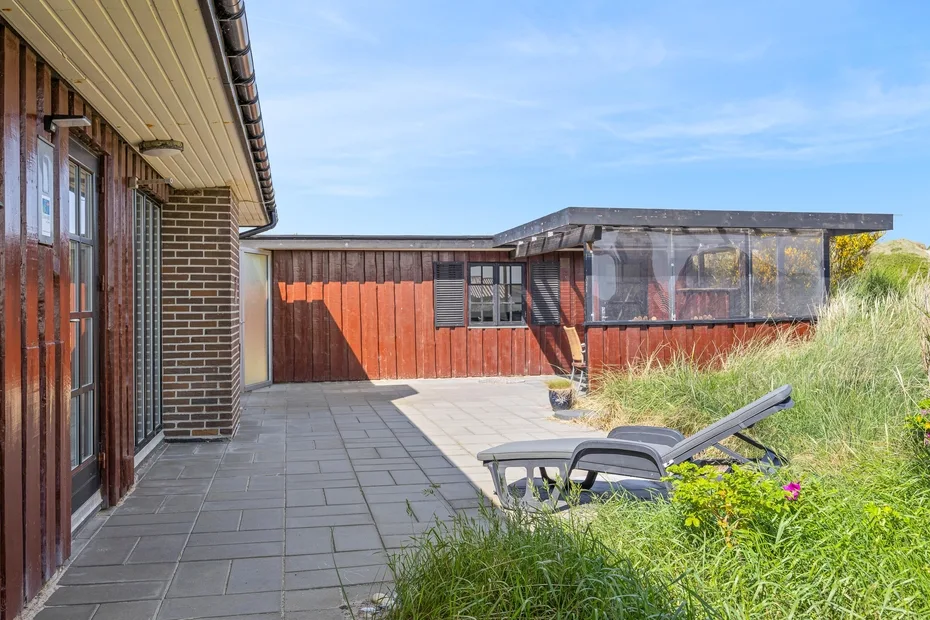 A paved outdoor patio beside a red wooden building, with lounge seating and tall grass.