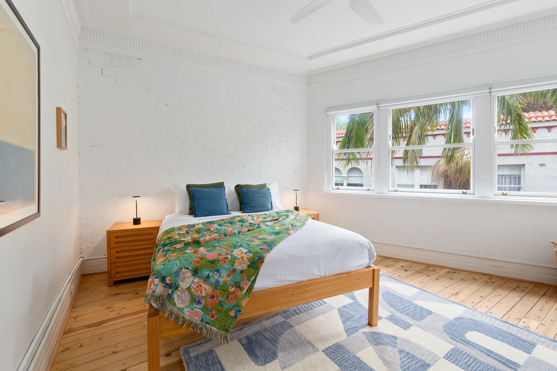 A bright bedroom with a bed covered in colorful floral bedding and windows overlooking greenery.
