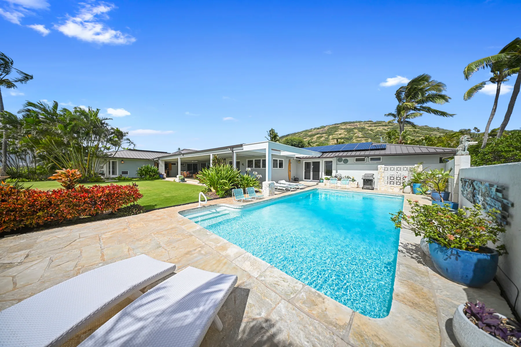 Outdoor pool beside a low-rise house, with lounge seating and tropical landscaping.