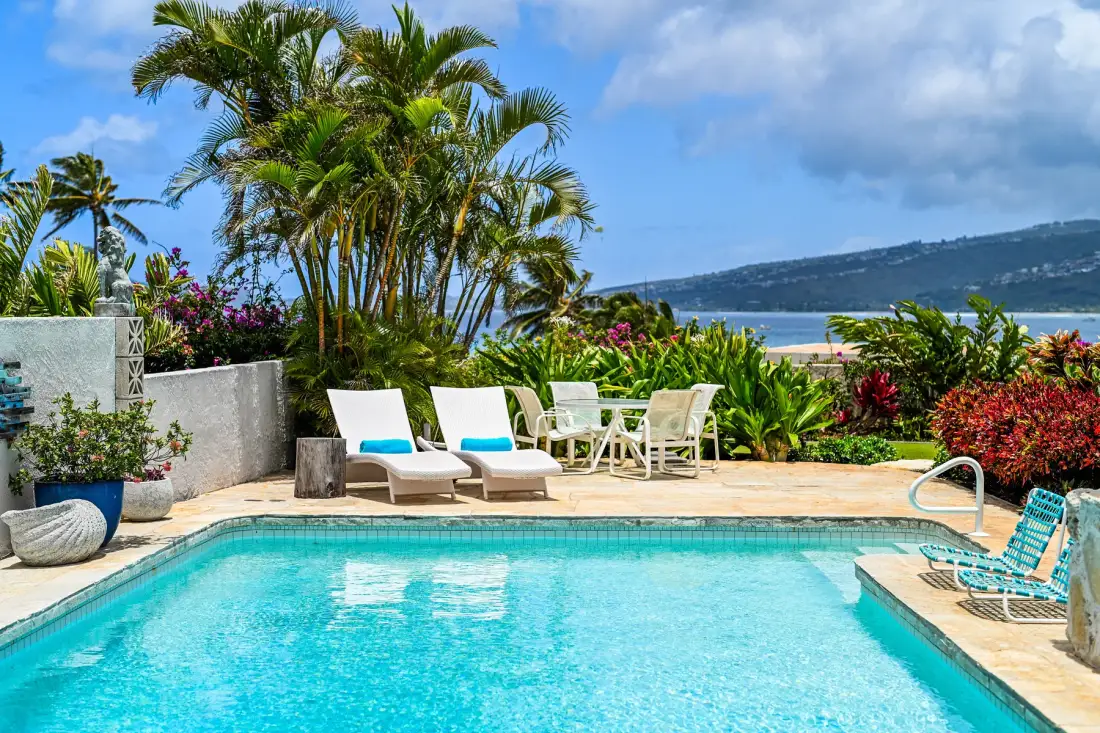 An outdoor swimming pool is surrounded by lounge seating and tropical plants.