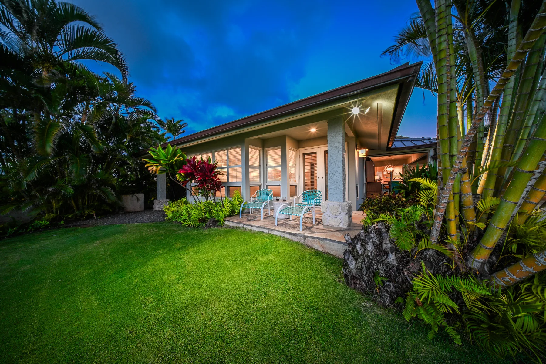 A modern house stands beside a green lawn, surrounded by tropical plants.