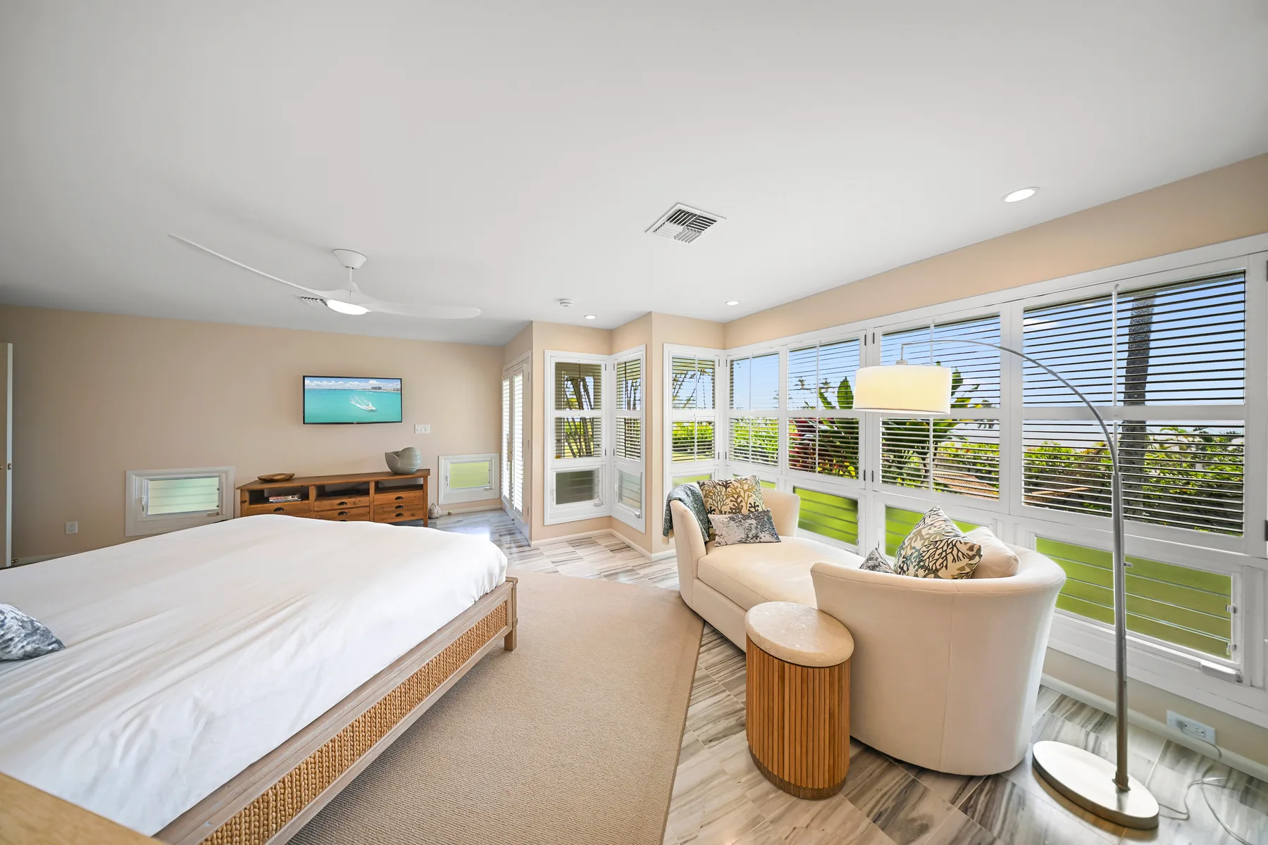 A bright bedroom with a bed, seating area, and broad windows overlooking greenery.