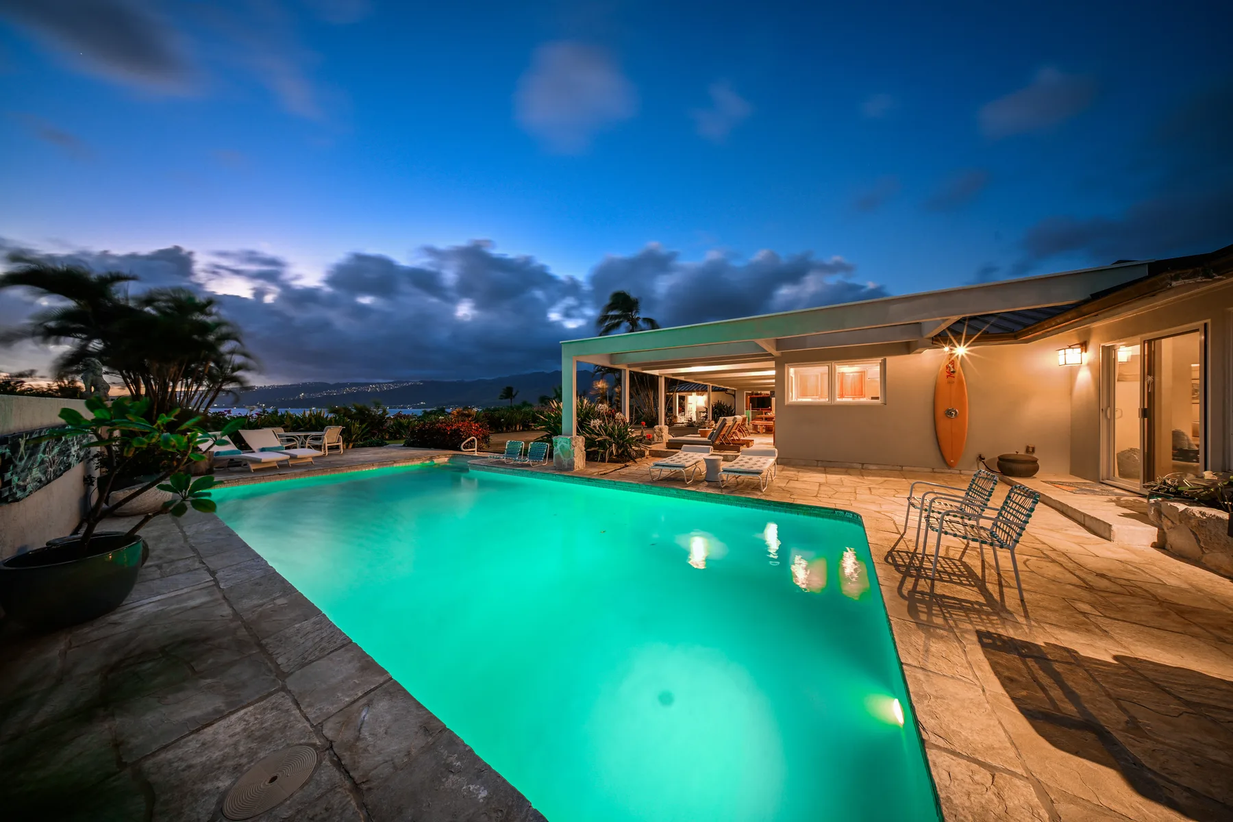 An illuminated swimming pool beside a low house and outdoor seating at dusk.