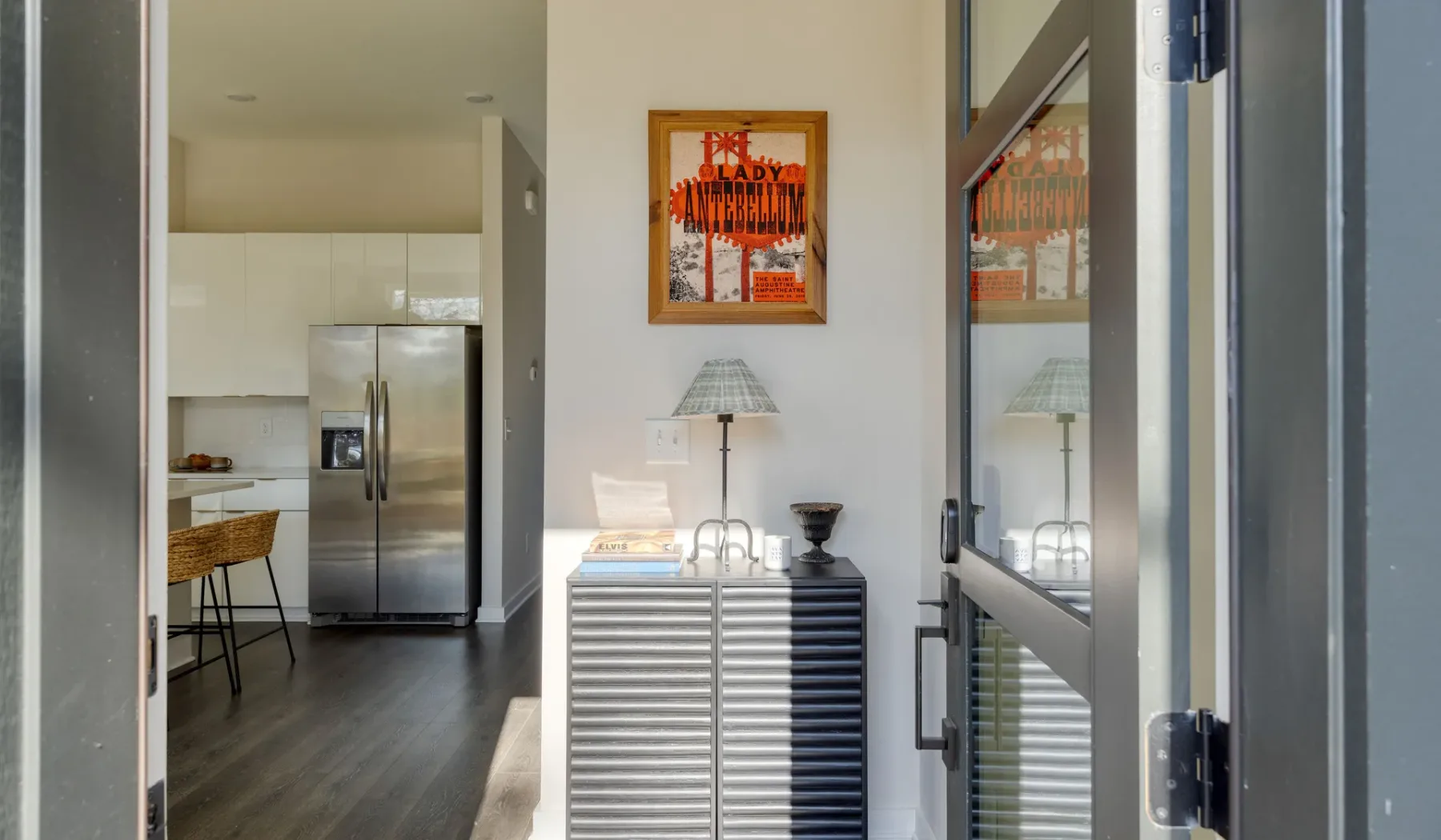 Una cocina luminosa y una entrada con un mueble metálico bajo un póster enmarcado.