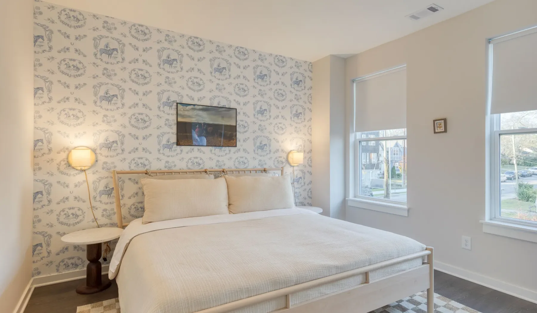 Un dormitorio con una cama tendida, papel tapiz estampado y dos ventanas.