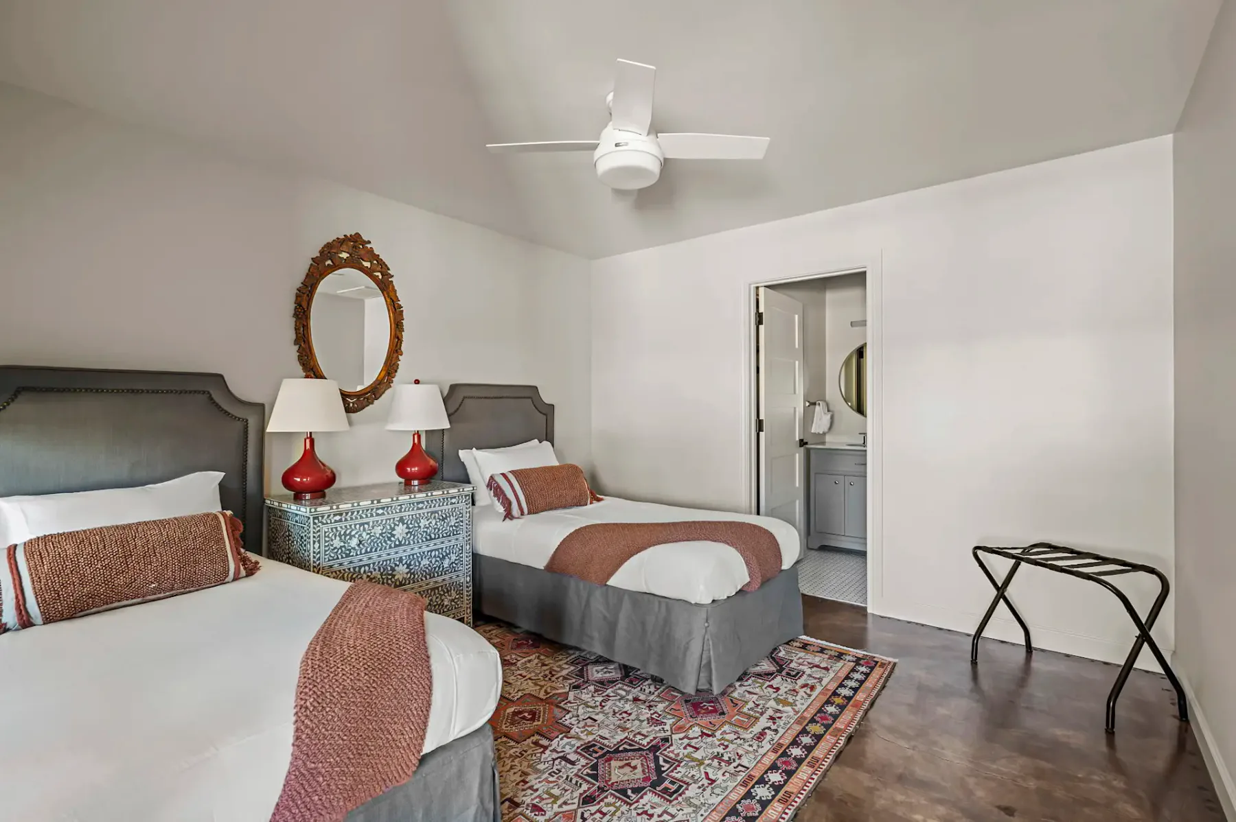 Dormitorio con dos camas individuales, una alfombra decorativa, lámparas rojas sobre una mesa auxiliar azul y un espejo redondo. Se ven un ventilador de techo y una puerta de baño.