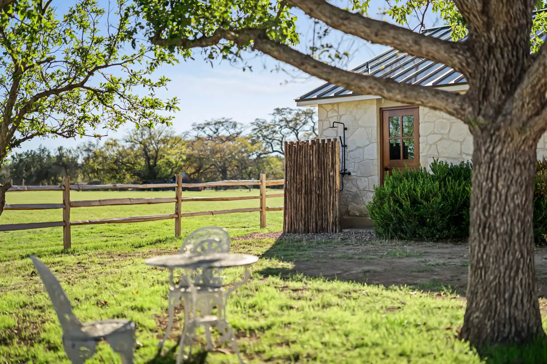 Casa de campo rústica de piedra con techo metálico, rodeada de una cerca de madera, árboles y una pequeña zona exterior con una mesa y una silla de metal blanco.